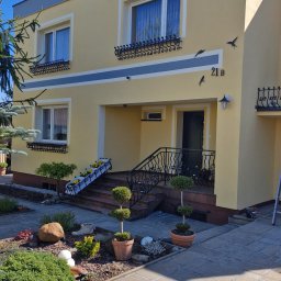 Sch 04 remonty - Elewacja domu po malowaniu w odcieniach żółci i szarości, z dekoracyjnymi elementami i numerem 21B. Uroku dodają metalowe ozdoby okien i rzeźby ptaków.