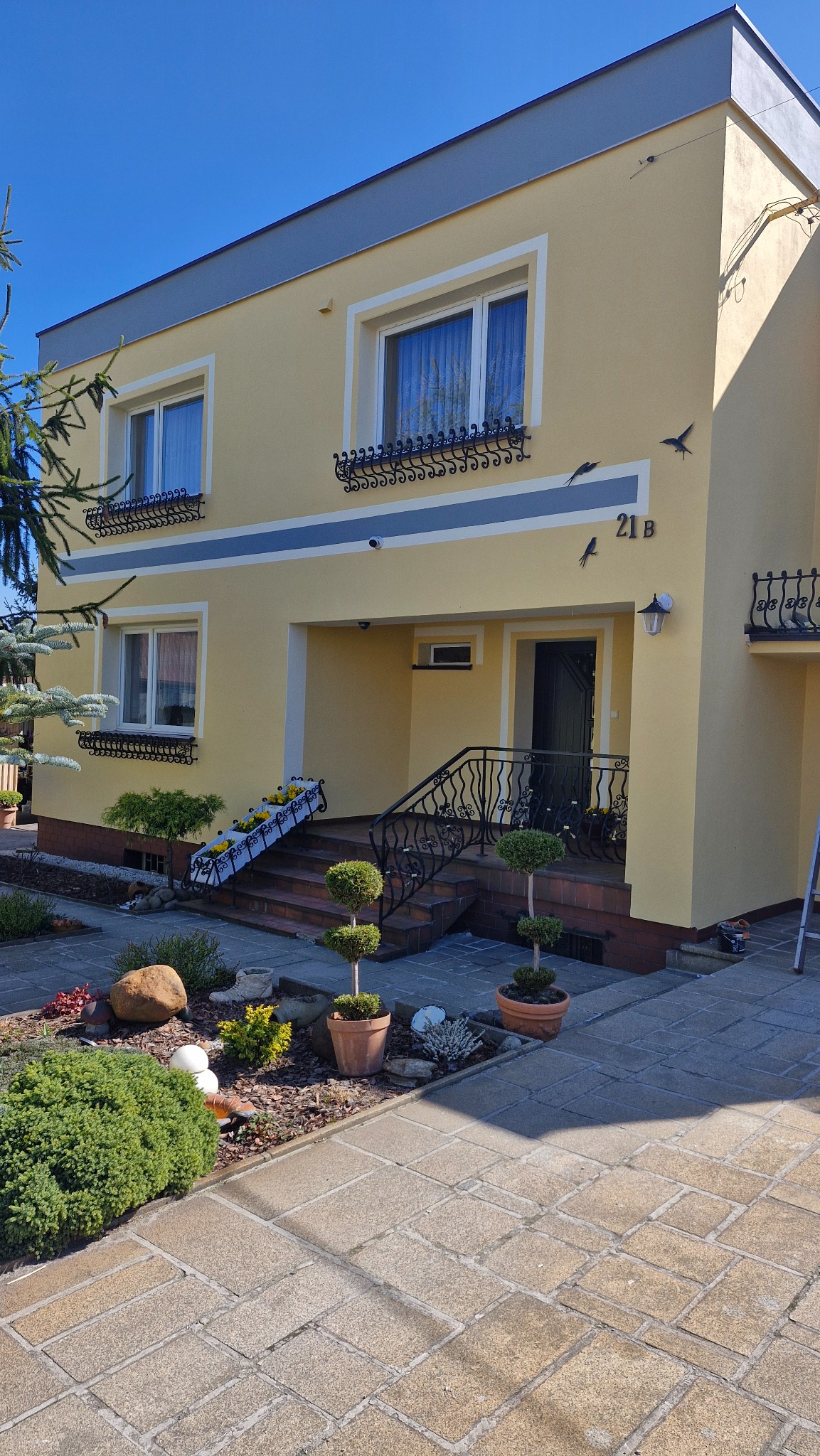 Elewacja domu po malowaniu w odcieniach żółci i szarości, z dekoracyjnymi elementami i numerem 21B. Uroku dodają metalowe ozdoby okien i rzeźby ptaków.