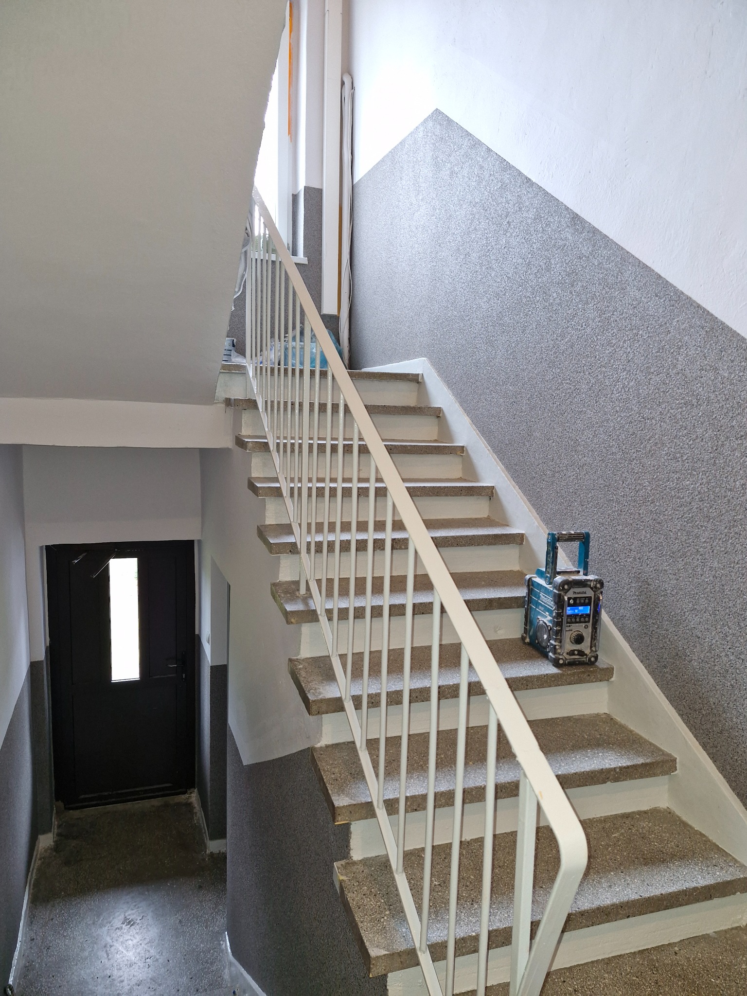Klatka schodowa po malowaniu: białe ściany, szary tynk strukturalny na ścianie bocznej. Na schodach radio budowlane Makita. Widok z dołu, w tle czarne drzwi.