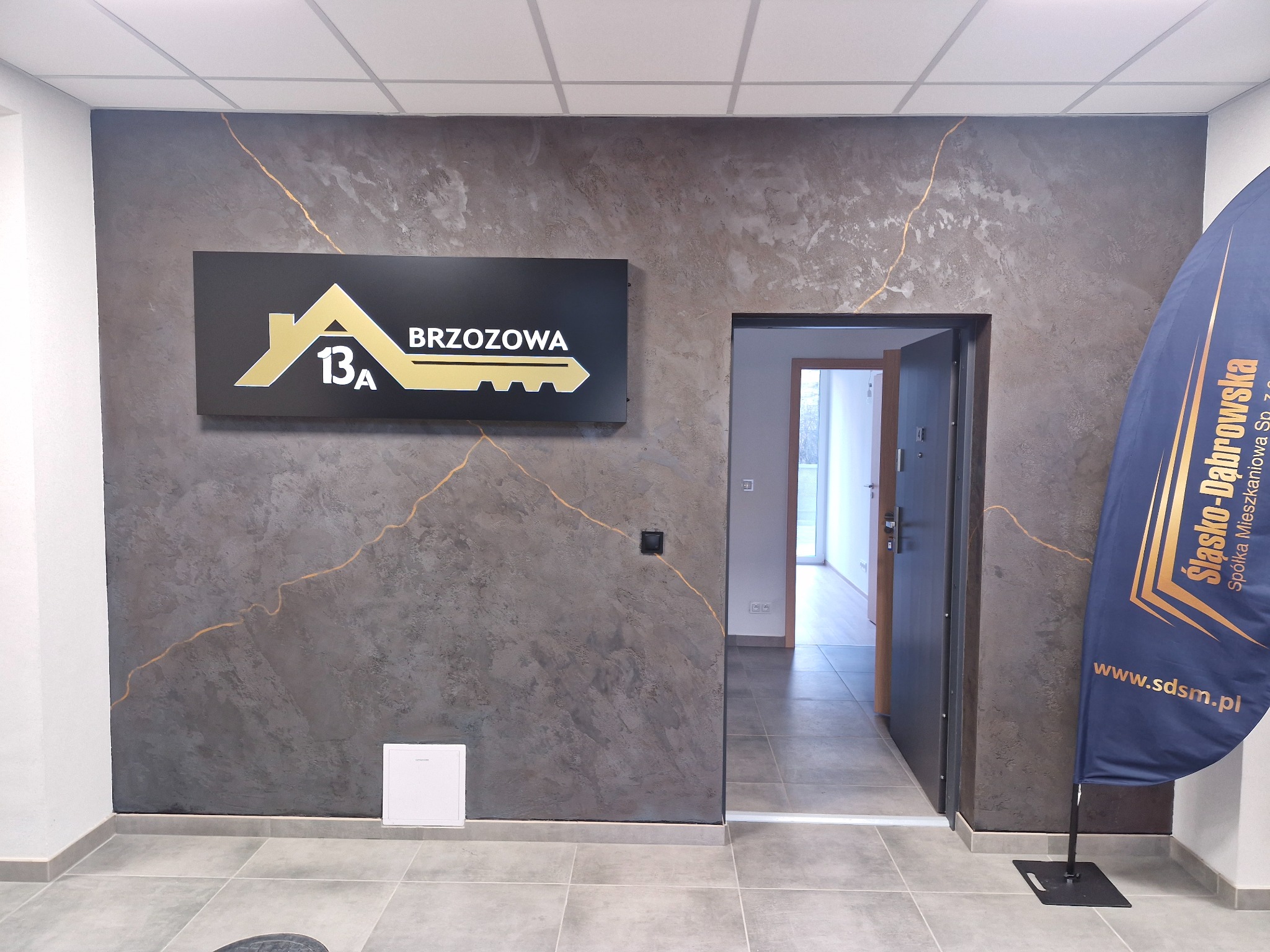Ściana z efektem dekoracyjnym w odcieniach szarości z subtelnymi złotymi żyłkami. Nad drzwiami szyld z adresem. W tle widoczne wnętrze biura.