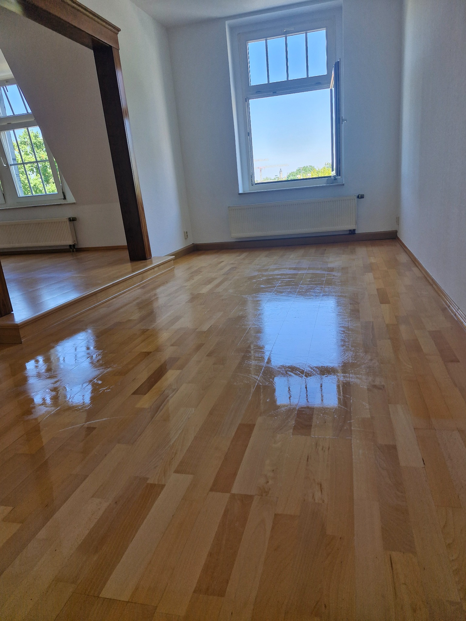 Jasne, lakierowane panele podłogowe w pokoju z oknem, wpadające światło odbija się od powierzchni, widoczne detale drewnianej konstrukcji.