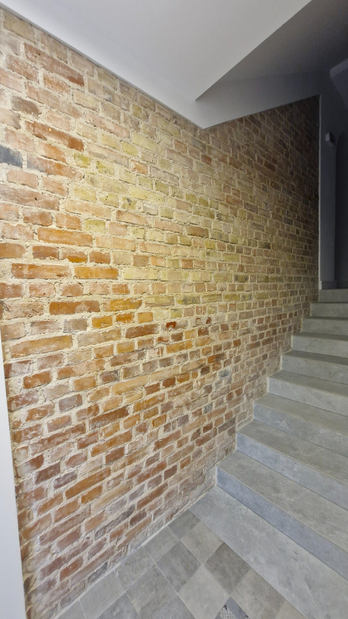 Ściana z cegły w odcieniach żółci i czerwieni, kontrastująca z szarymi płytkami podłogowymi i schodami. Biały sufit dopełnia wnętrze. Nietypowe połączenie materiałów.