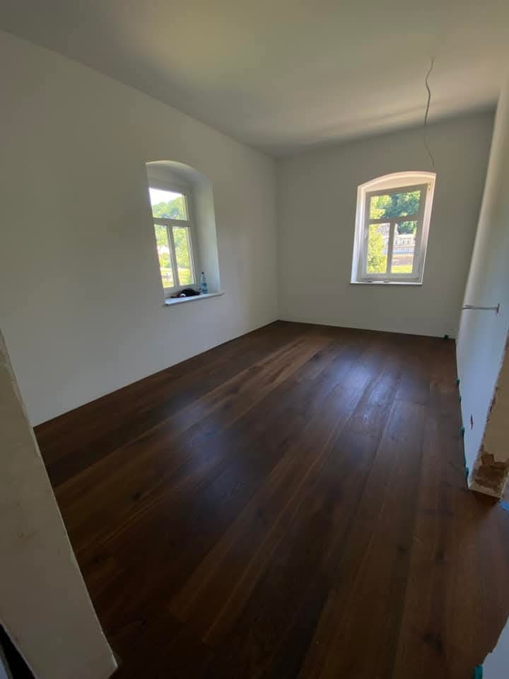 Pokój po remoncie: świeżo pomalowane białe ściany, ciemna drewniana podłoga, dwa okna z widokiem na zieleń. Kabel zwisa z sufitu, ściana częściowo nieotynkowana.