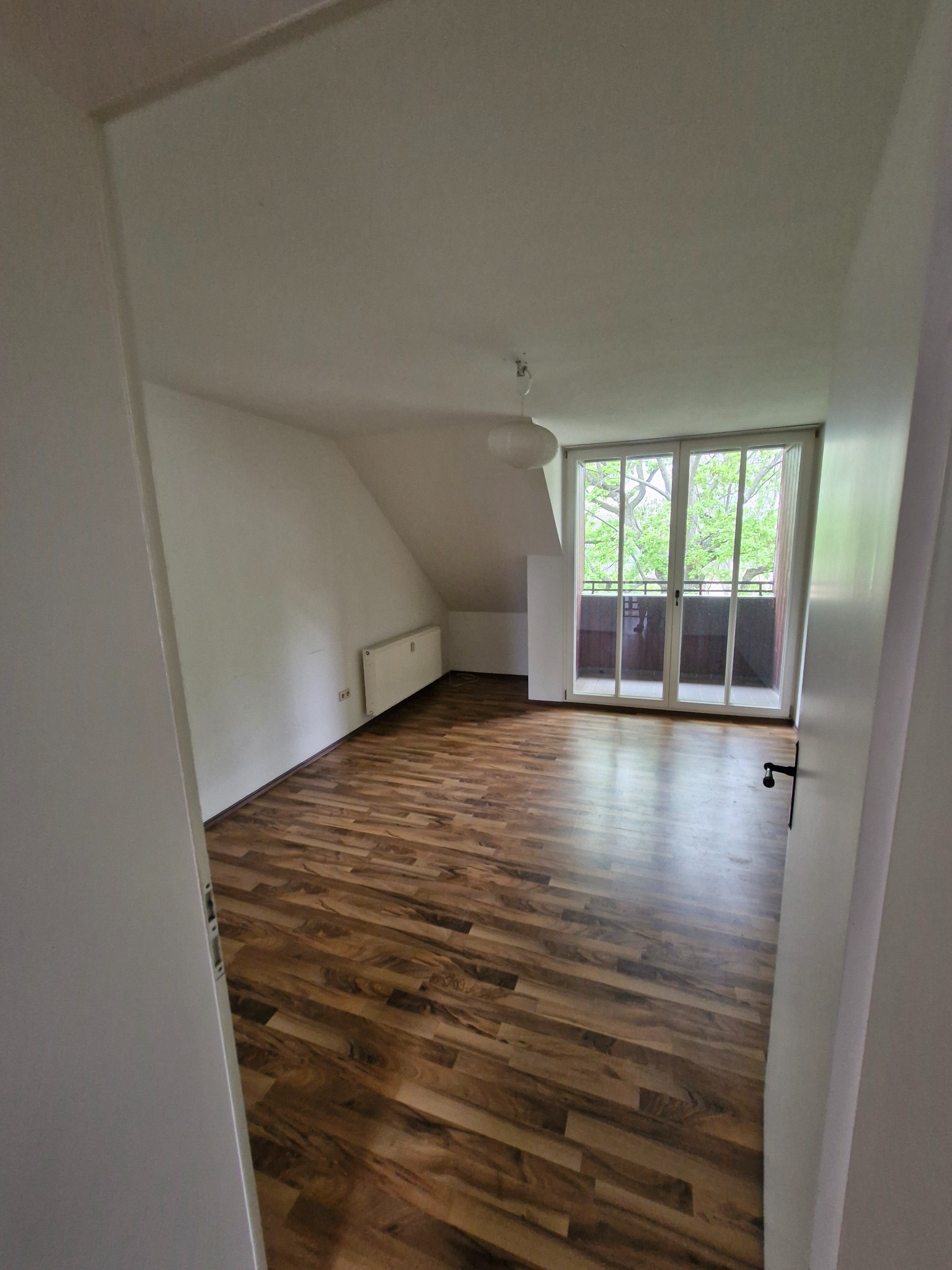 Poddasze z drewnianą podłogą i balkonem. Ściany białe, widoczny grzejnik. Ujęcie zza otwartych drzwi, z perspektywy wnętrza. Jasne, naturalne światło.