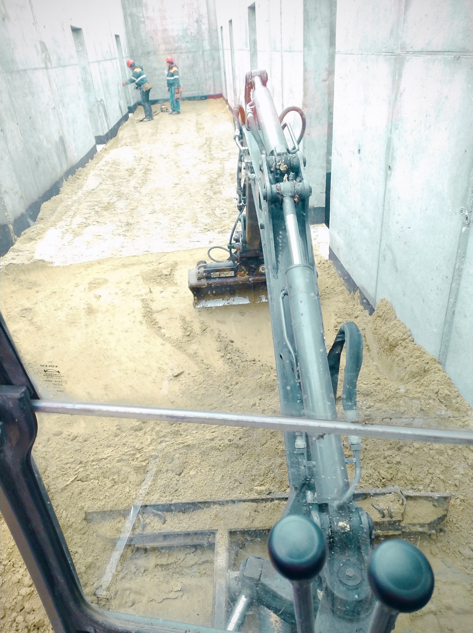 Widok z kabiny mini koparki na utwardzany piaskiem wykop pod fundamenty budynku. W tle dwóch robotników w kaskach.