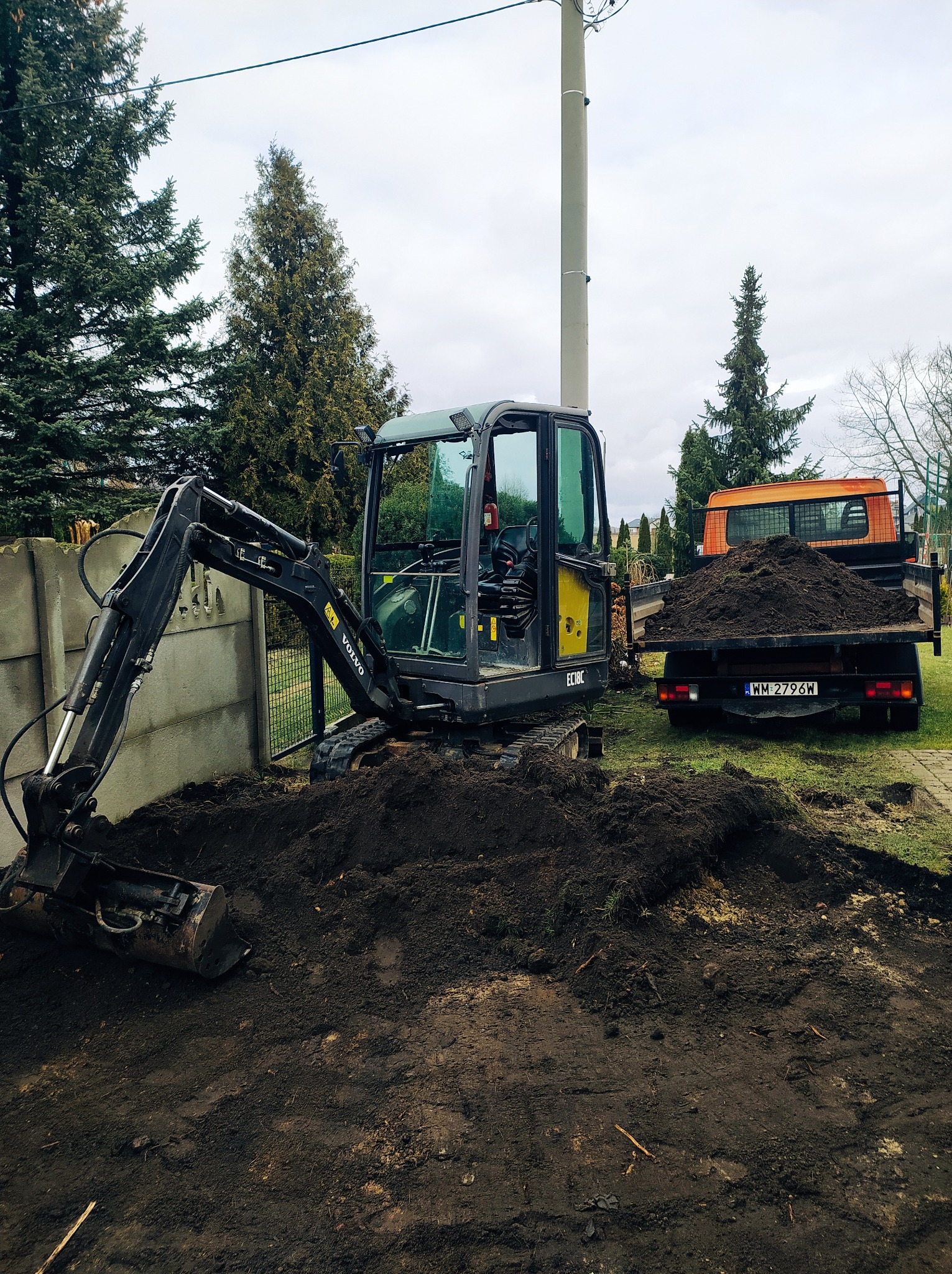 Czarny mini-koparka Volvo EC18C na działce z wykopaną ziemią, obok stoi pomarańczowa wywrotka z ziemią. W tle widoczne drzewa i betonowe ogrodzenie.