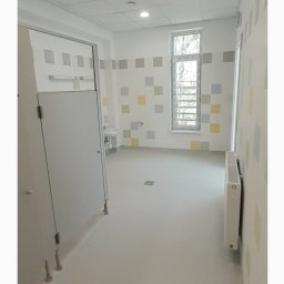 Lubelskie Domy - Wnętrze wykończonej łazienki publicznej z kabinami, umywalką i grzejnikiem. Ściany zdobią kwadratowe płytki w pastelowych kolorach. Podłoga jasna, antypoślizgowa.