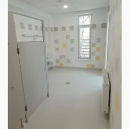 Wnętrze wykończonej łazienki publicznej z kabinami, umywalką i grzejnikiem. Ściany zdobią kwadratowe płytki w pastelowych kolorach. Podłoga jasna, antypoślizgowa.