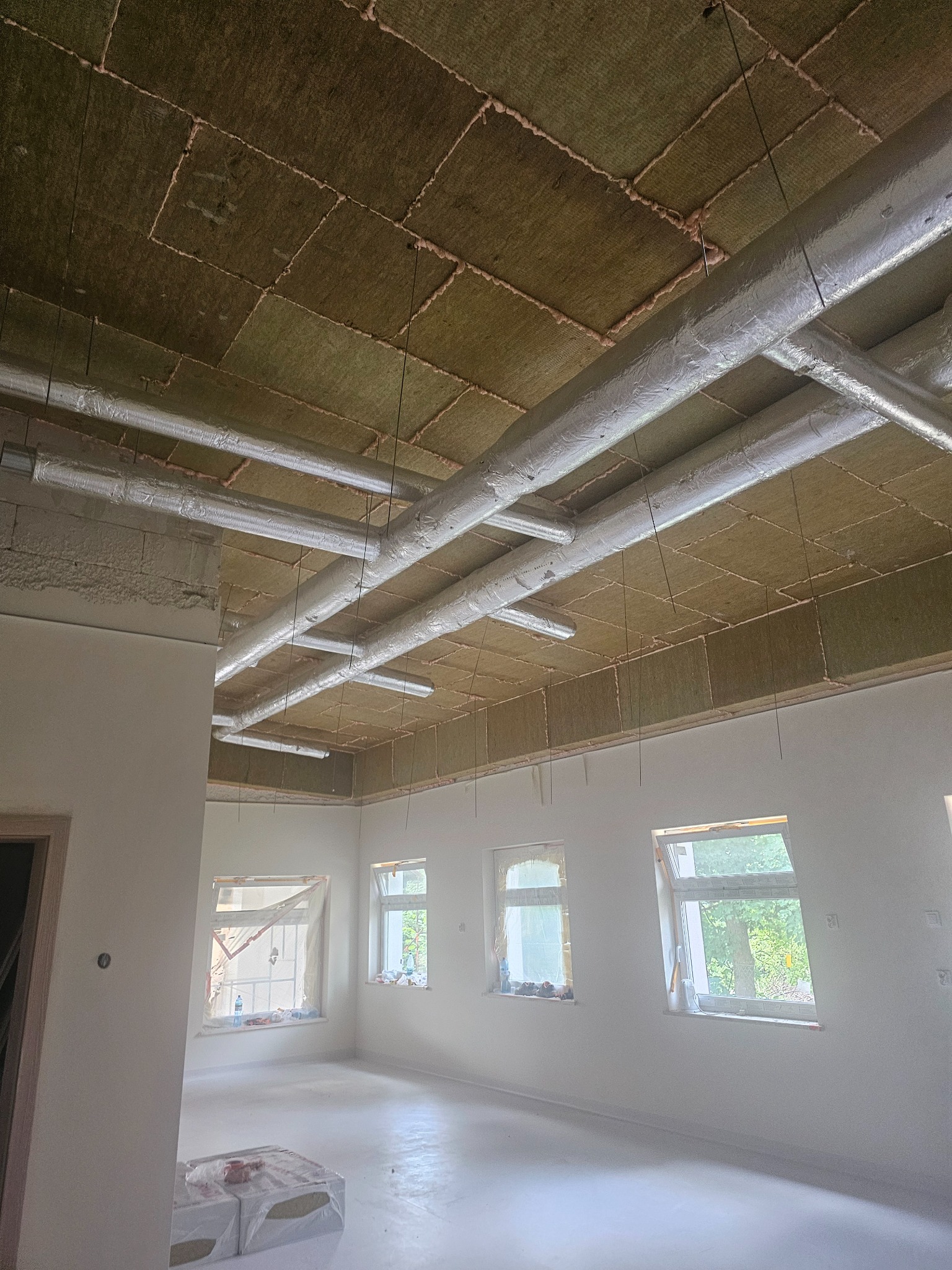 Ocieplony sufit wełną mineralną i instalacją wentylacyjną w nowym, jasnym pomieszczeniu z oknami. Widoczne detale montażu i izolacji.