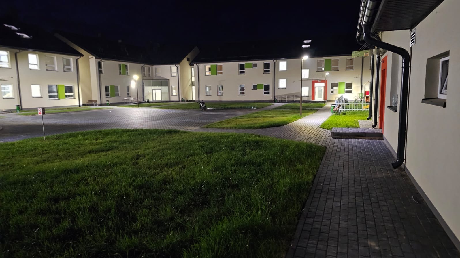 Nowoczesny budynek z zielonymi akcentami okiennymi i czerwonymi drzwiami, oświetlony nocą, z trawnikiem i chodnikiem z kostki brukowej na pierwszym planie.