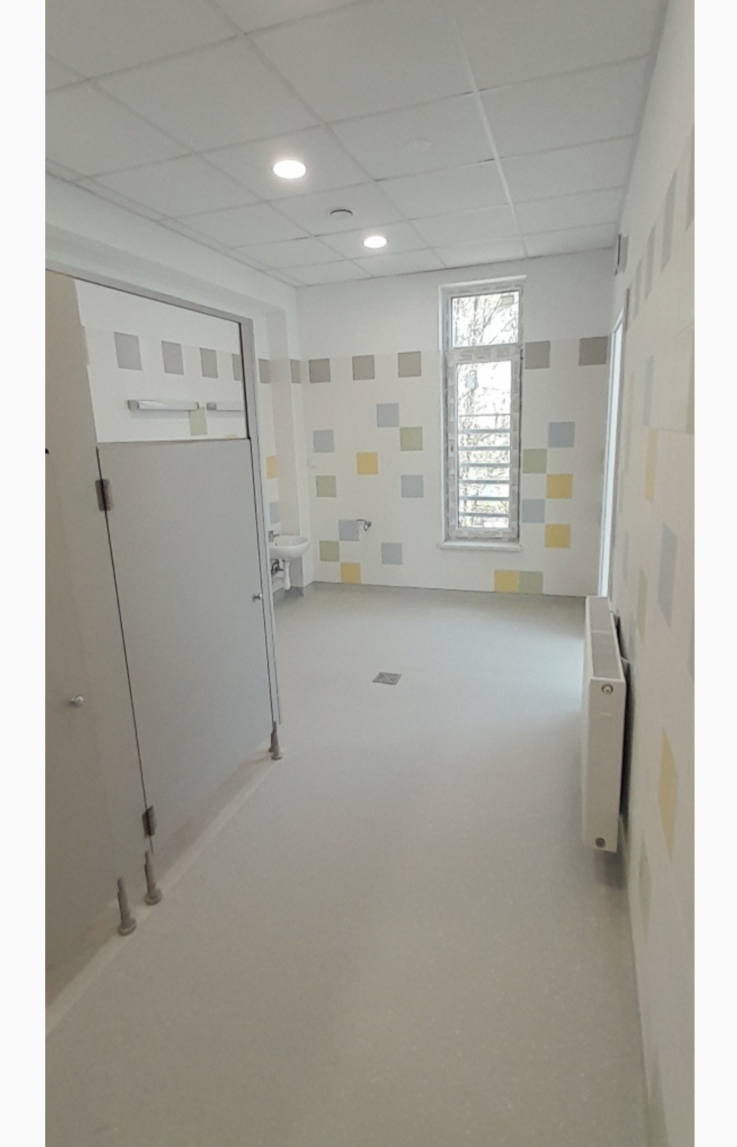 Wnętrze wykończonej łazienki publicznej z kabinami, umywalką i grzejnikiem. Ściany zdobią kwadratowe płytki w pastelowych kolorach. Podłoga jasna, antypoślizgowa.