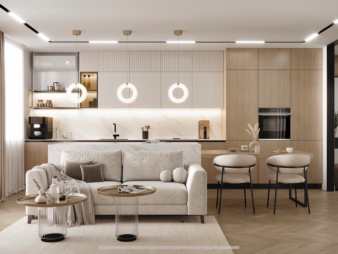 Beżowa kuchnia z wyspą połączona z salonem. Nad wyspą wiszą okrągłe lampy. W tle drewniane szafki i sprzęt AGD. Na pierwszym planie sofa i stoliki kawowe.