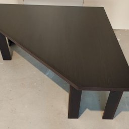 HANDEL OBWOŹNY I STACJONARNY MEBLAMI DARIUSZ IWAŃSKI - Ciemny, drewniany stół o nietypowym kształcie, z grubym blatem i solidnymi nogami, stoi na szarej podłodze. W tle biała ściana z gniazdkiem.