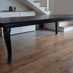 HANDEL OBWOŹNY I STACJONARNY MEBLAMI DARIUSZ IWAŃSKI - Elegancki, czarny stół z giętymi nogami na drewnianej podłodze. W tle biała komoda i schody. Widoczna część okna z widokiem na zieleń.