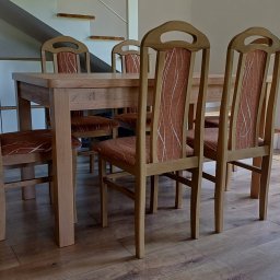 HANDEL OBWOŹNY I STACJONARNY MEBLAMI DARIUSZ IWAŃSKI - Drewniany stół jadalniany z krzesłami o charakterystycznych, wysokich oparciach i brązowych siedziskach, na tle jasnego wnętrza z drewnianą podłogą i schodami.