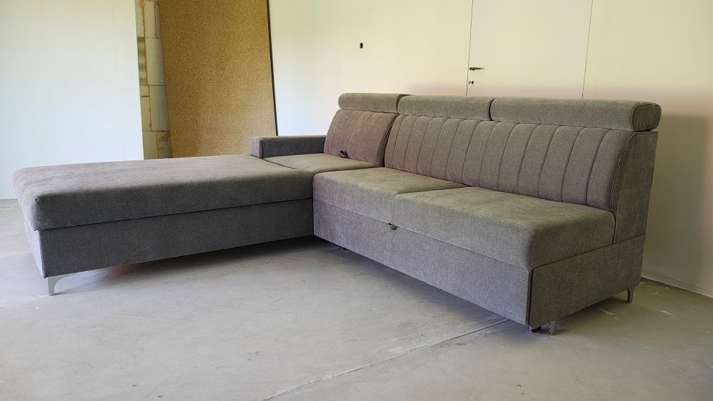 Szara, rozkładana sofa narożna z funkcją spania i pojemnikiem na pościel, na metalowych nóżkach, na tle surowej ściany i podłogi.