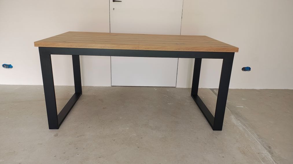 Stół z drewnianym blatem i czarnymi, metalowymi nogami w surowym wnętrzu. Widoczne niedokończone ściany i podłoga, dodające industrialnego charakteru.