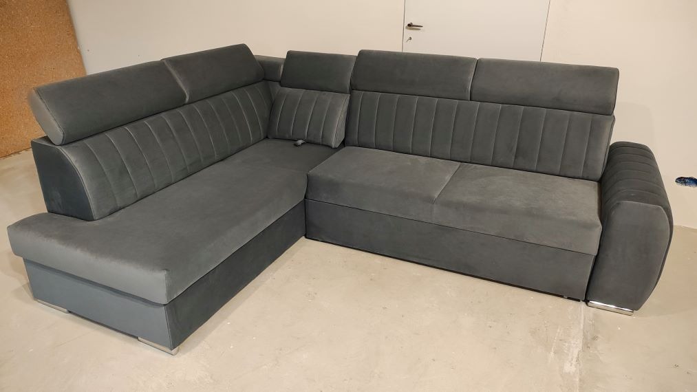 Szara, tapicerowana sofa narożna z regulowanymi zagłówkami i poduszką, ustawiona na jasnej podłodze. Widoczne metalowe nóżki i pionowe przeszycia na oparciu.