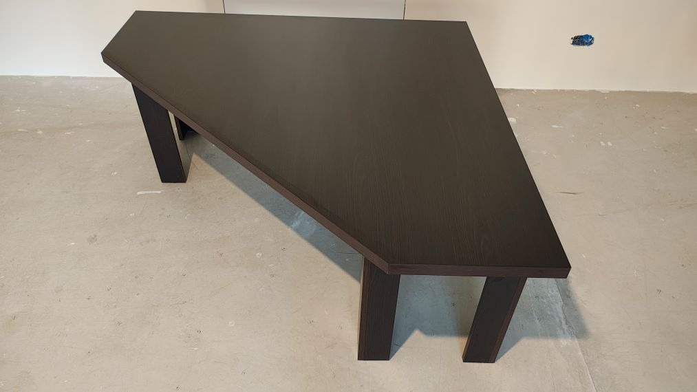 Ciemny, drewniany stół o nietypowym kształcie, z grubym blatem i solidnymi nogami, stoi na szarej podłodze. W tle biała ściana z gniazdkiem.