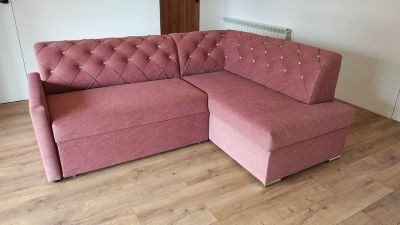 Różowa, tapicerowana sofa narożna z pikowanym oparciem i dekoracyjnymi guzikami, na drewnianej podłodze. Widoczny fragment ściany i drzwi.