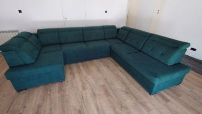 Szmaragdowy narożnik tapicerowany w kształcie litery U z poduszkami na oparciu, na jasnej podłodze z paneli, z widocznym grzejnikiem i fragmentem białej ściany.