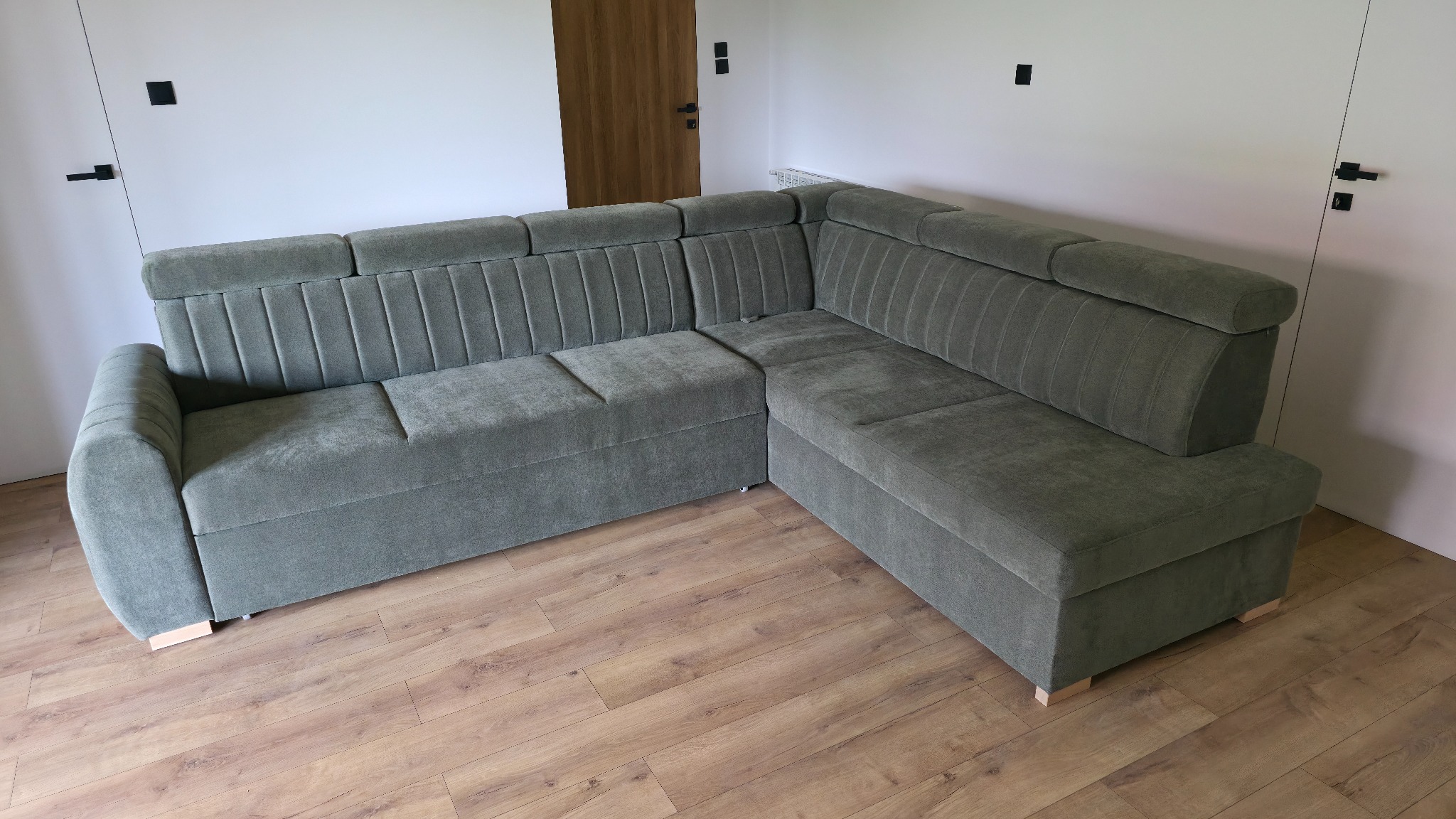 Szary, welurowy narożnik z pionowymi przeszyciami na oparciu, ustawiony w rogu jasnego pokoju z drewnianą podłogą i białymi drzwiami.