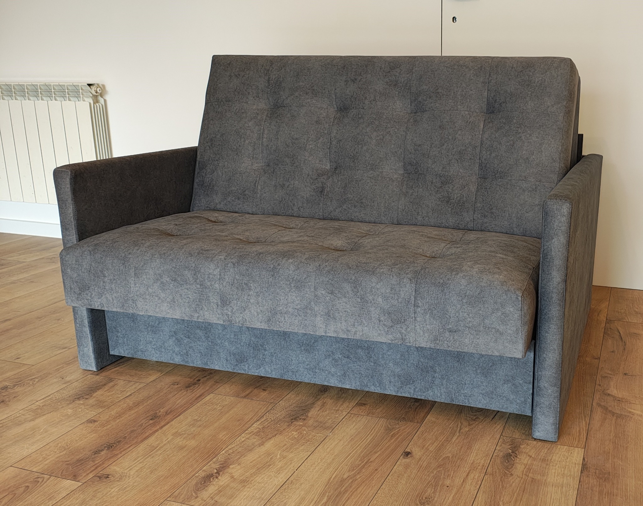 Szara, tapicerowana sofa dwuosobowa z pikowanym oparciem, na drewnianej podłodze, z widocznym grzejnikiem w tle. Nowoczesny design i neutralna kolorystyka.