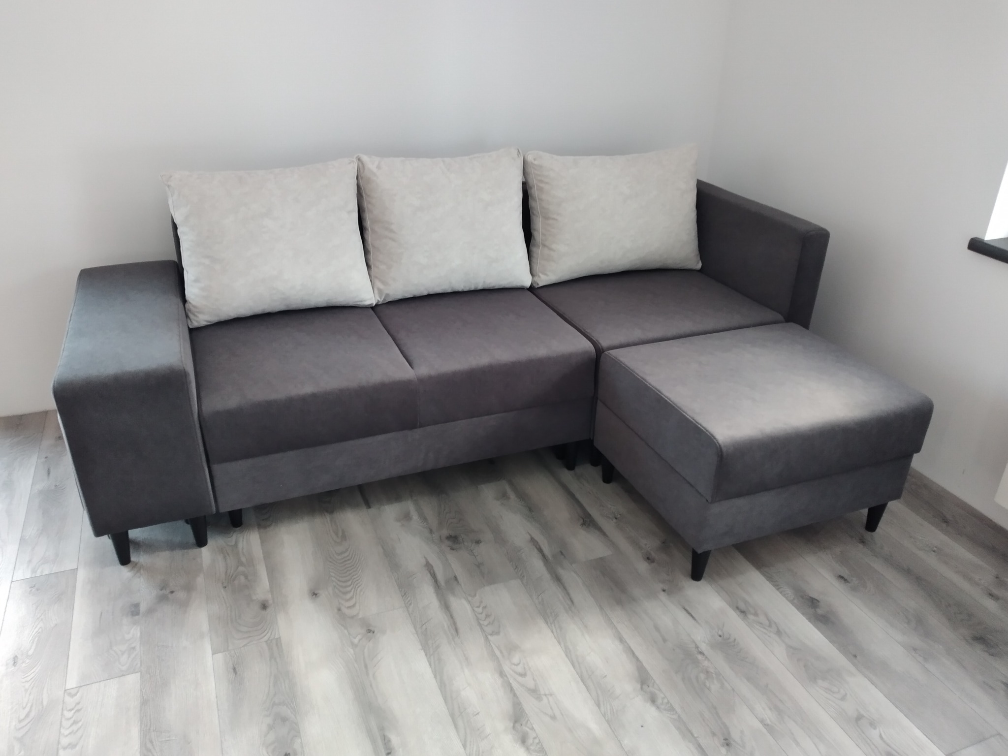 Szary narożnik z trzema poduszkami w jasnym odcieniu, z dodatkowym pufem, na podłodze z paneli imitujących drewno, minimalistyczny styl.