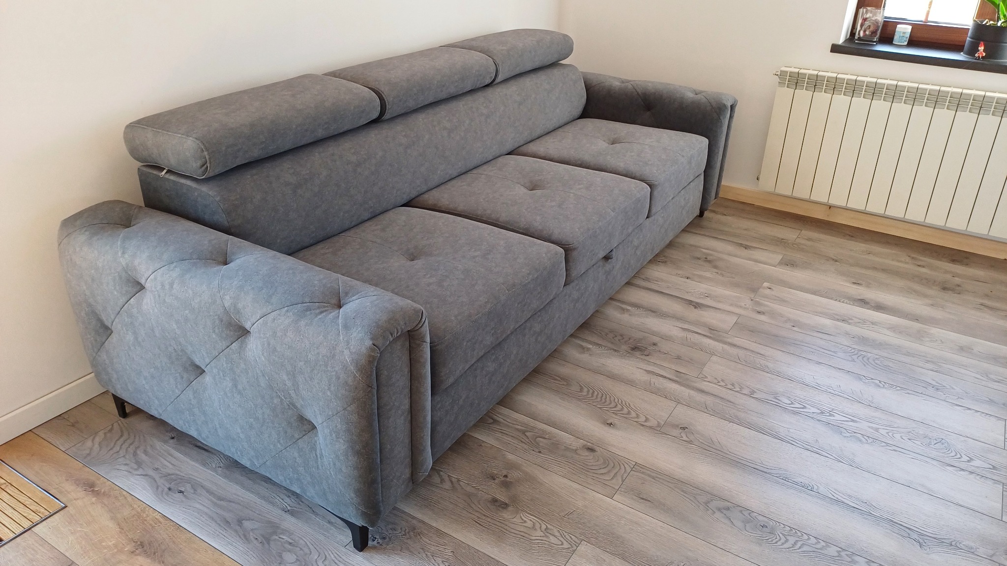 Szara, tapicerowana sofa z regulowanymi zagłówkami i pikowanymi bokami, ustawiona na drewnianej podłodze w jasnym wnętrzu.