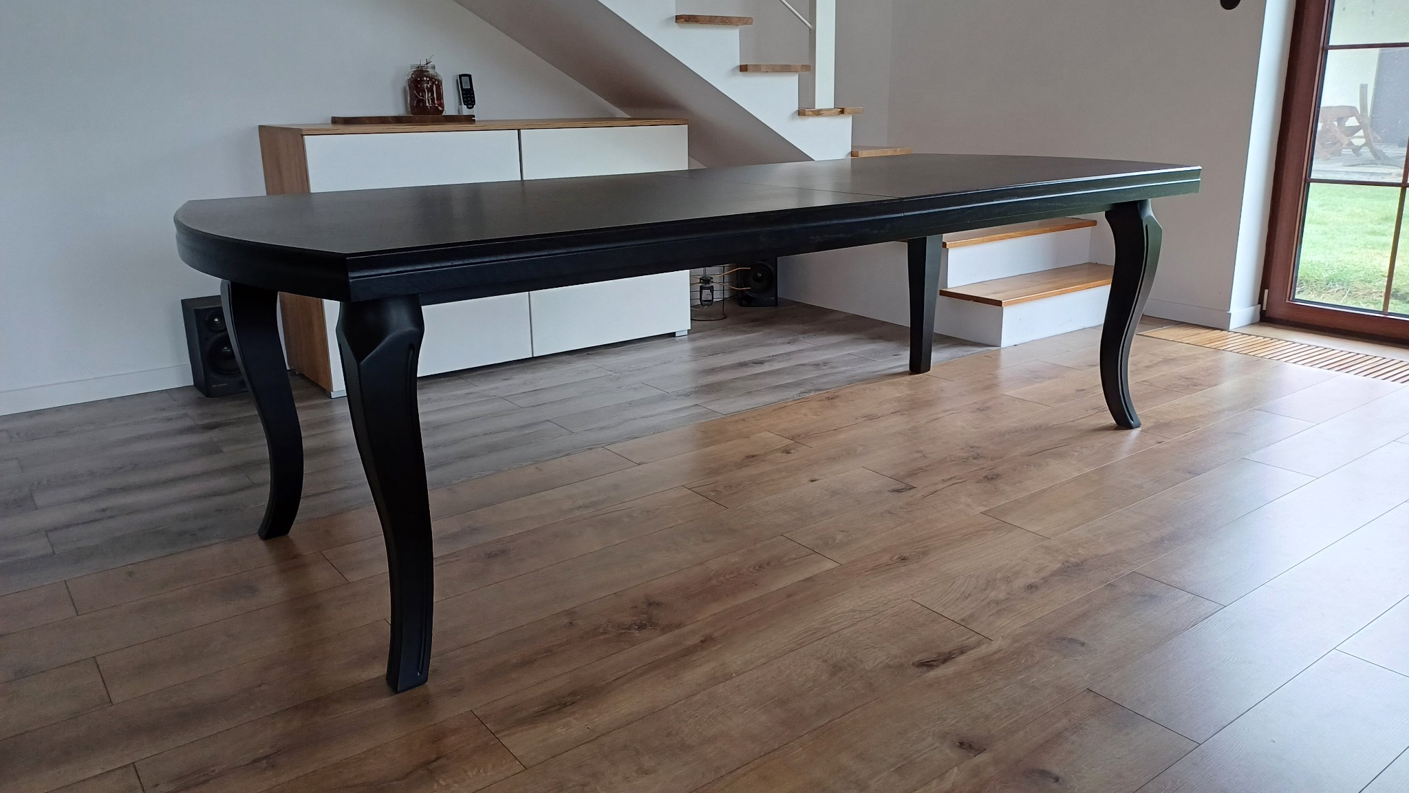 Elegancki, czarny stół z giętymi nogami na drewnianej podłodze. W tle biała komoda i schody. Widoczna część okna z widokiem na zieleń.