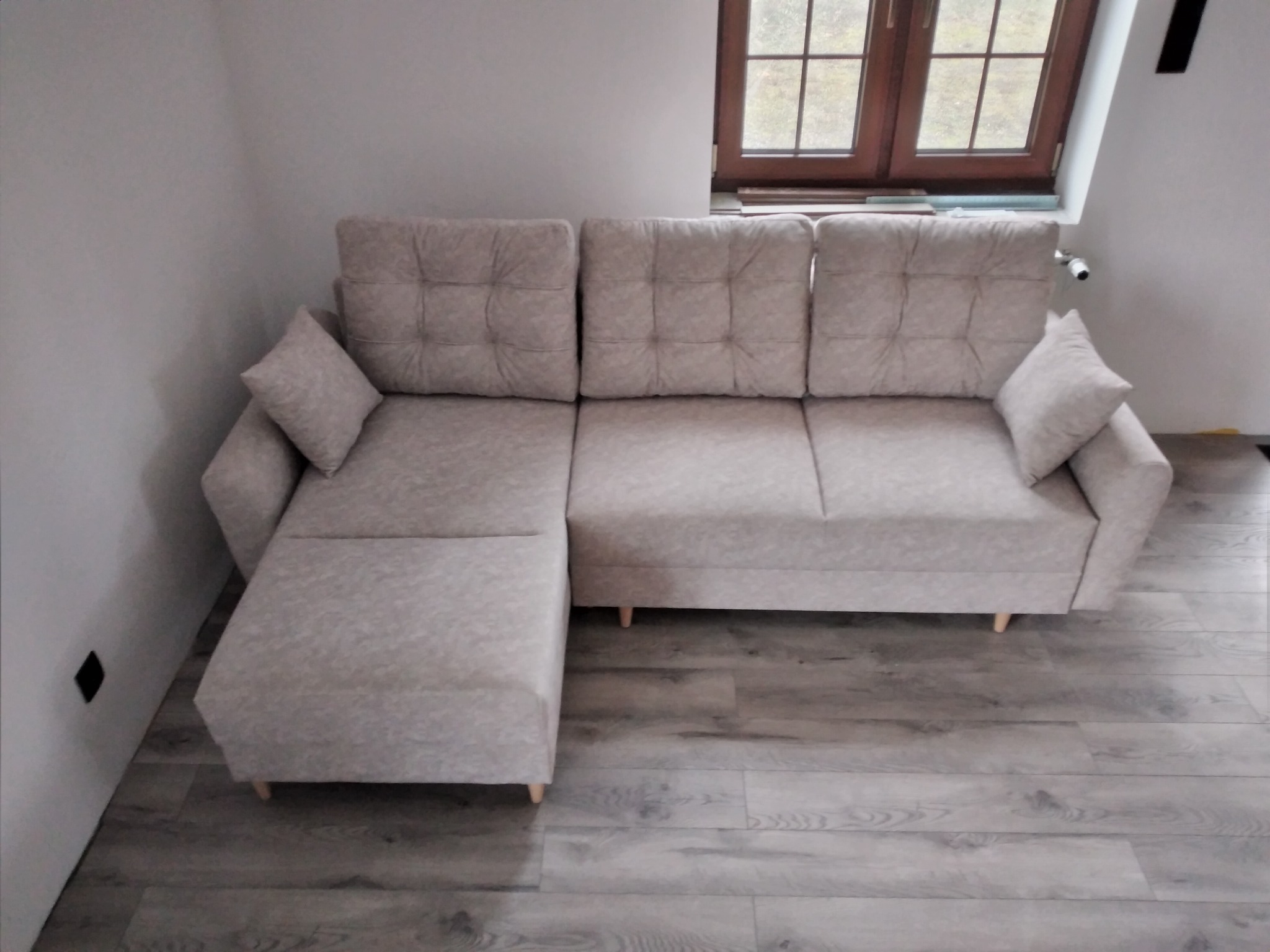 Jasnoszara sofa narożna z pikowanymi poduszkami oparcia, ustawiona na podłodze z paneli w odcieniach szarości, z oknem w tle.