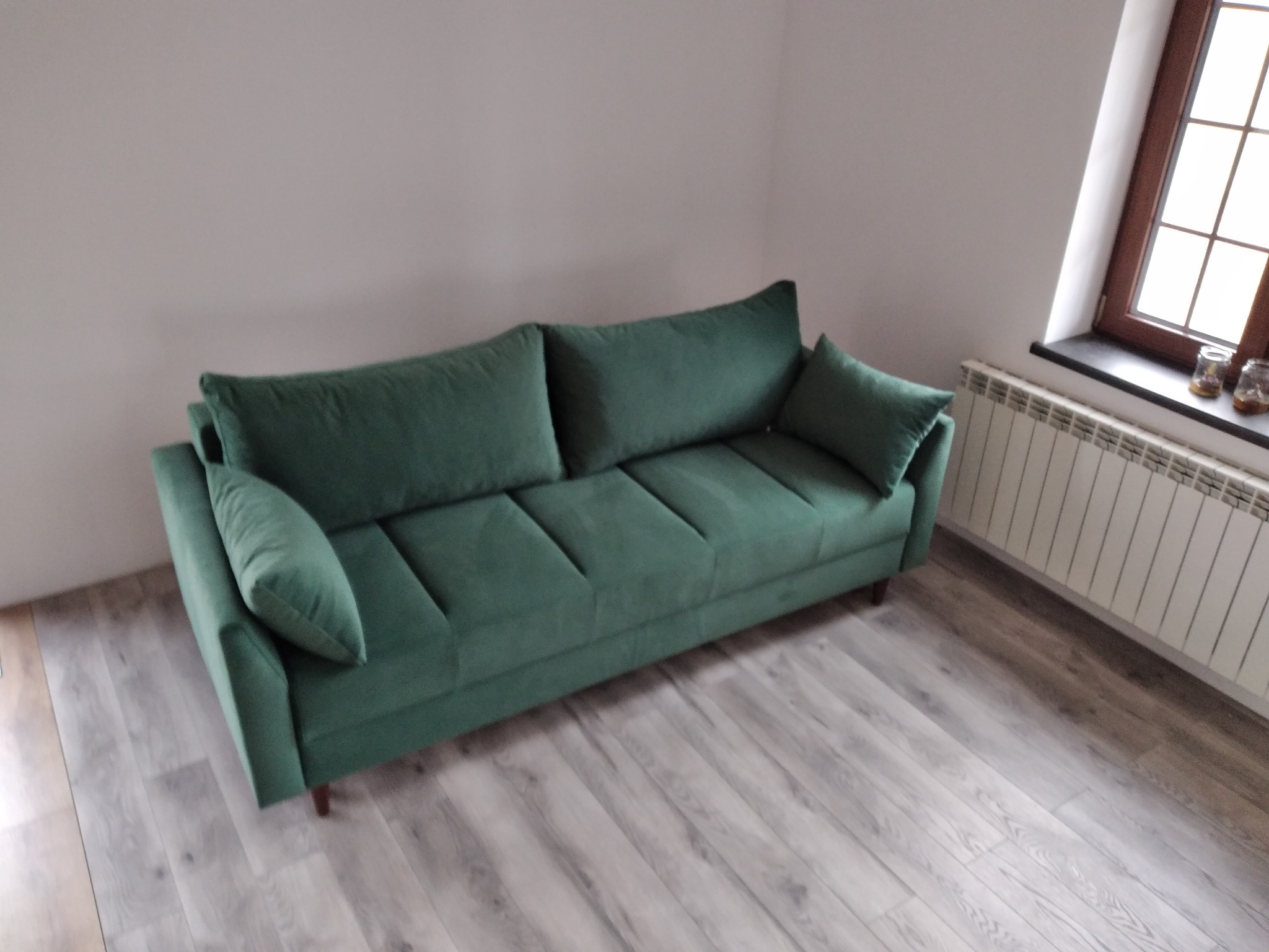Szmaragdowa sofa tapicerowana z poduszkami w salonie z drewnianą podłogą i oknem z grzejnikiem. Minimalistyczny design.