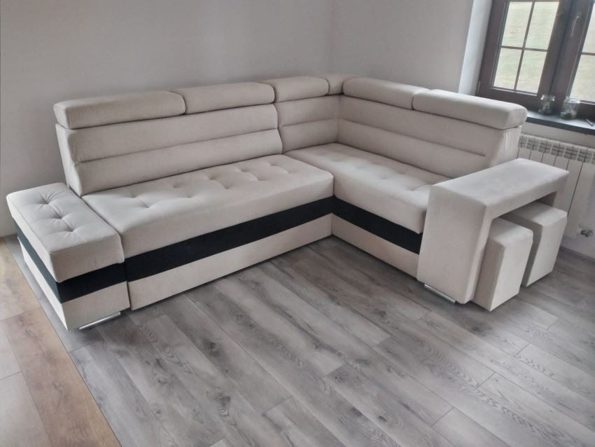 Jasny, narożny zestaw wypoczynkowy z poduchami i czarnym pasem, uzupełniony o dwa pufy. Całość na tle paneli podłogowych i okna.