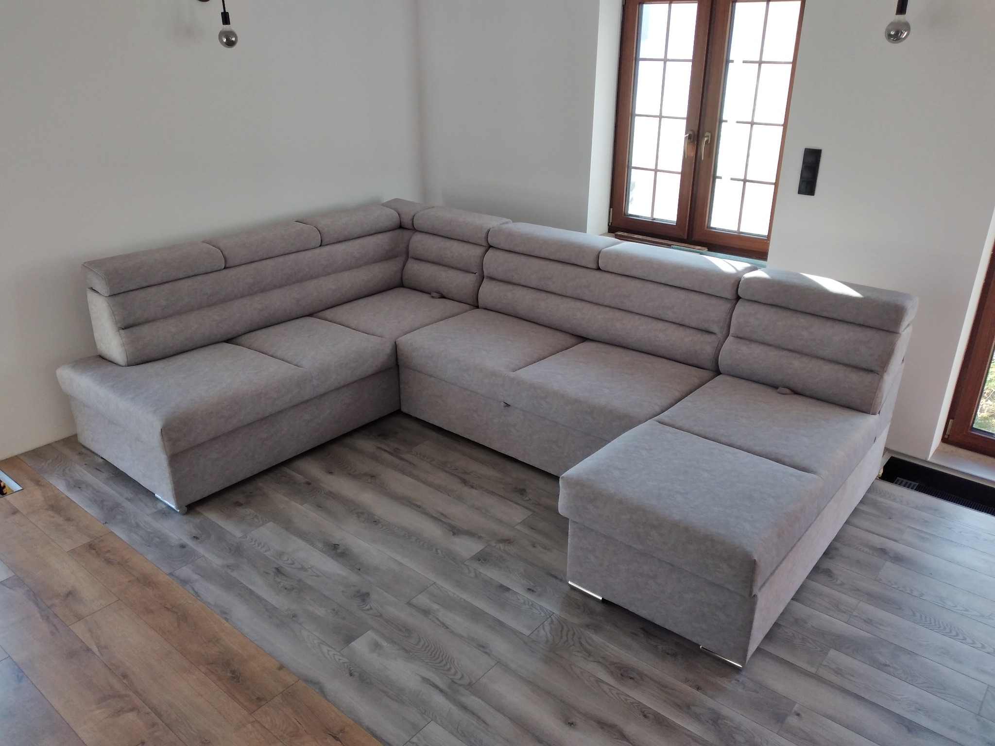 Jasnoszary, tapicerowany narożnik modułowy z poziomymi przeszyciami w nowoczesnym salonie z drewnianą podłogą i oknem. Minimalistyczne oświetlenie sufitowe.