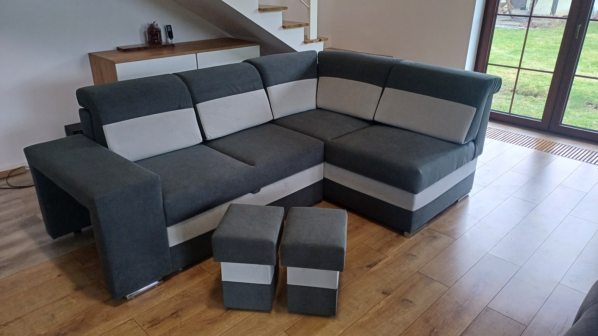 Szary narożnik z pufami, dwukolorowy, z jasnymi wstawkami, na drewnianej podłodze, w tle schody i okno z widokiem na zieleń. Nowoczesny design.
