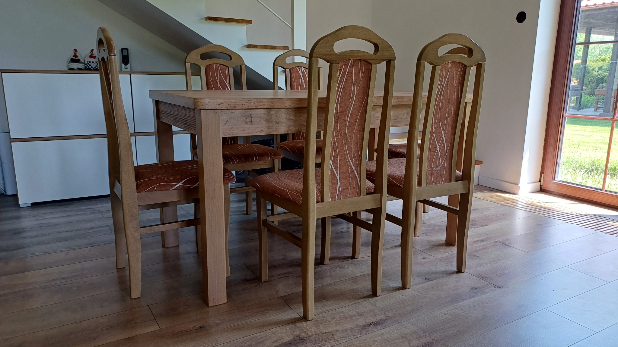 Drewniany stół jadalniany z krzesłami o charakterystycznych, wysokich oparciach i brązowych siedziskach, na tle jasnego wnętrza z drewnianą podłogą i schodami.