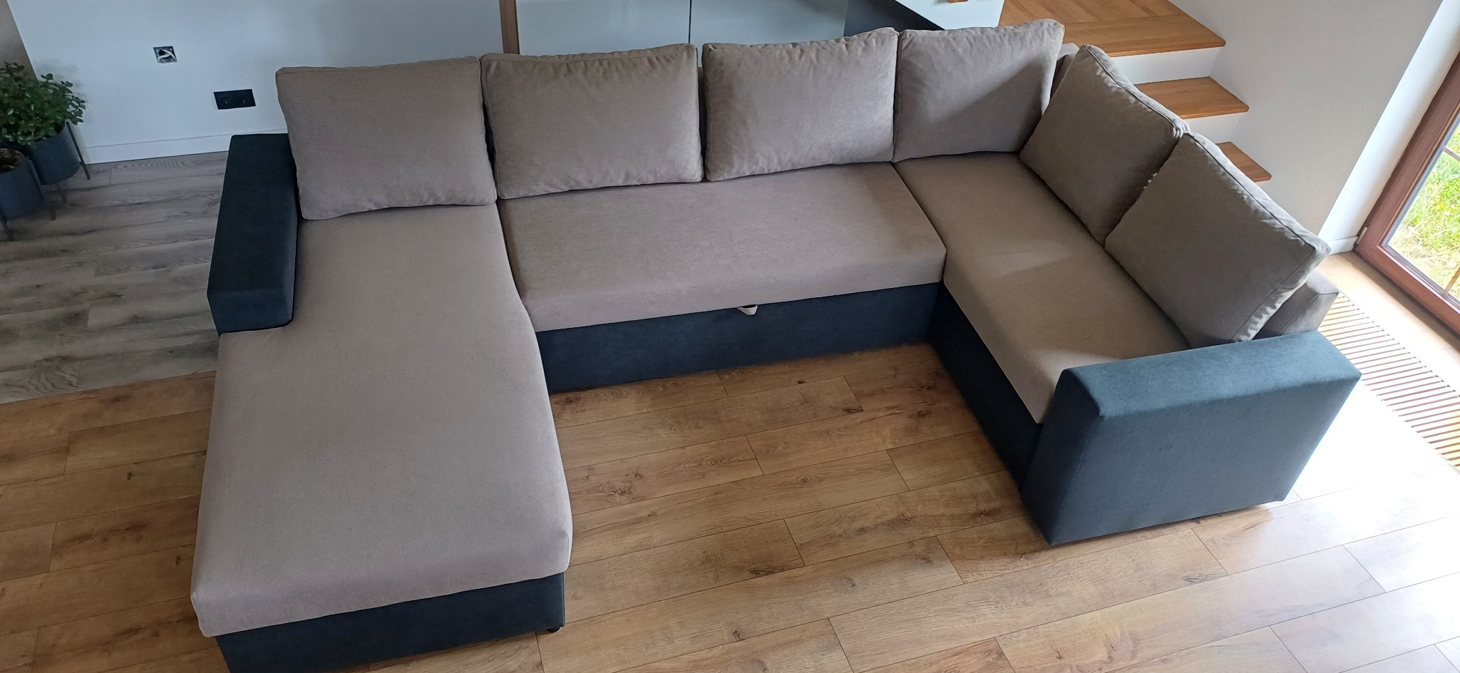 Narożnik tapicerowany w kształcie litery U, z szarymi bokami i beżowymi poduchami, na drewnianej podłodze, w tle schody i okno.