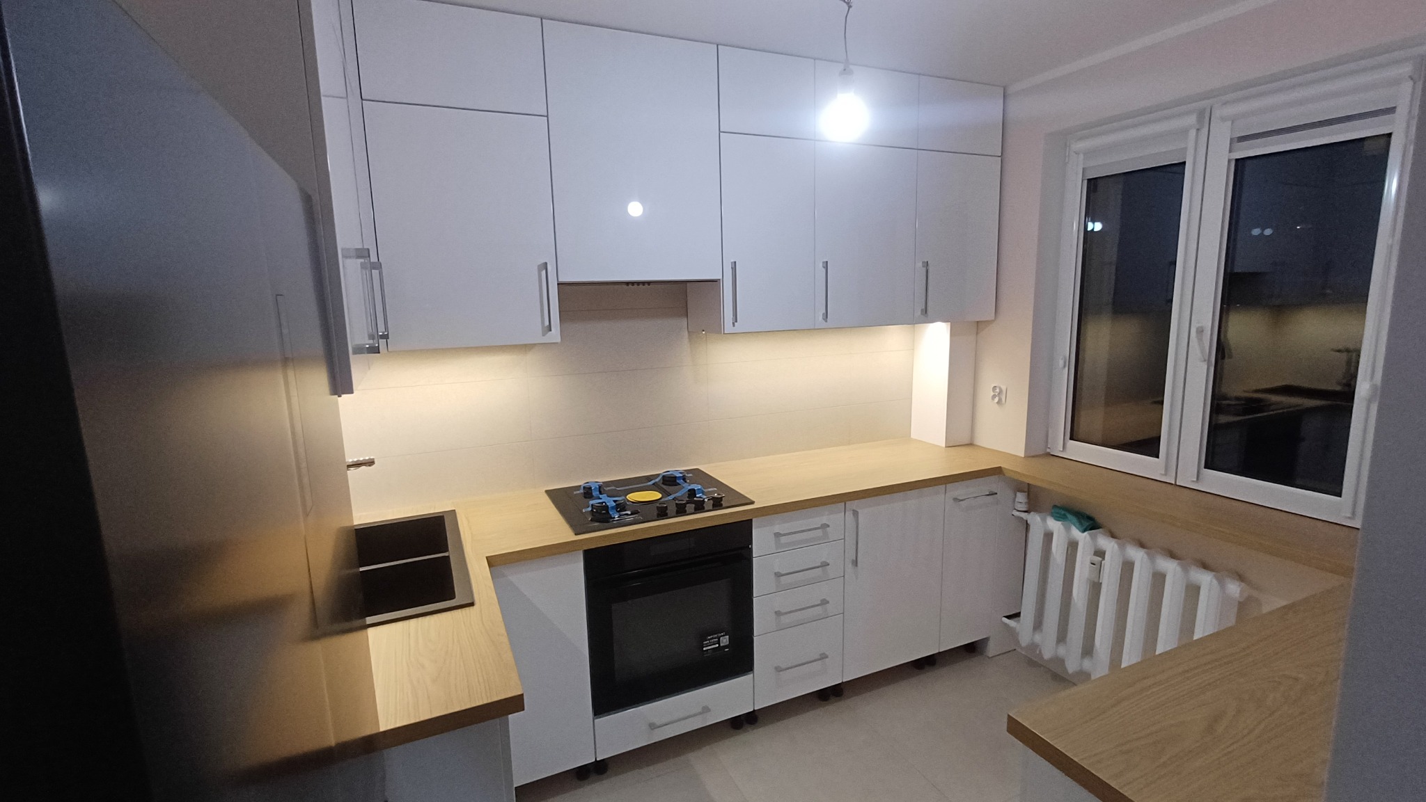 Biała kuchnia z drewnianym blatem i czarną płytą grzewczą. Nad blatem wiszące szafki. W tle okno z widokiem na zewnątrz. Minimalistyczny design.