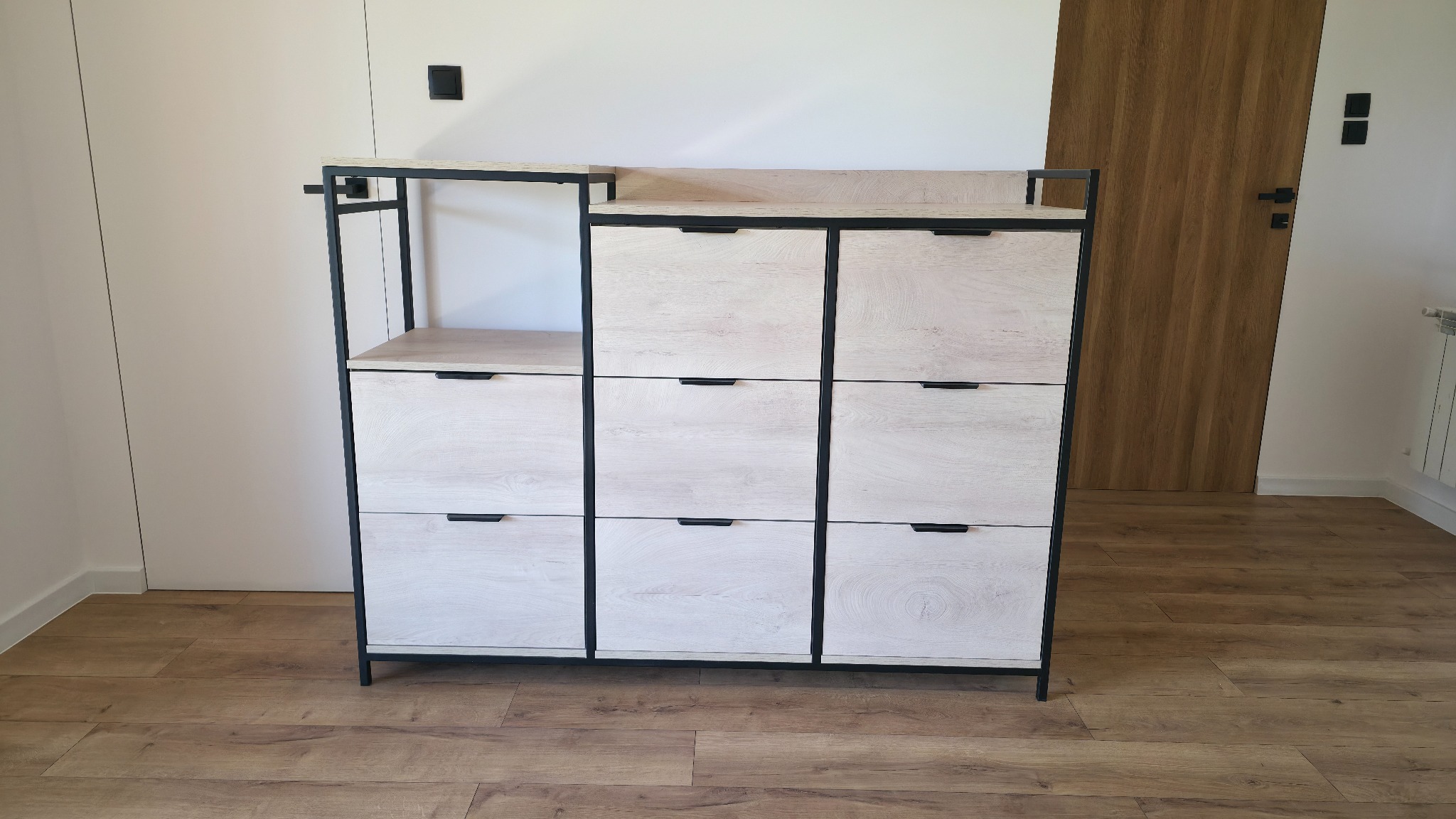 Nowoczesna szafka na buty z jasnego drewna i czarnych metalowych elementów, Wadowice. Minimalistyczny design, trzy rzędy szuflad i otwarte półki.