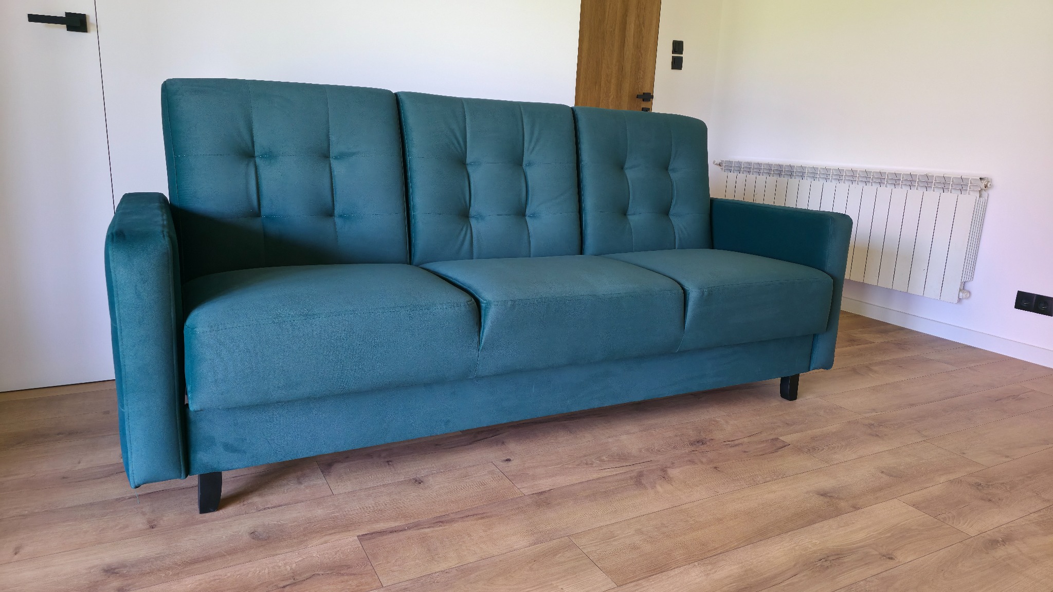 Turkusowa sofa trzyosobowa z pikowanym oparciem, ustawiona na drewnianej podłodze, na tle białej ściany z grzejnikiem. Minimalistyczny design, nowoczesne wnętrze.