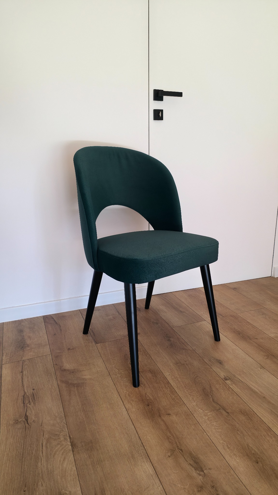 Stylowe, ciemnozielone krzesło tapicerowane z czarnymi nogami na drewnianej podłodze, na tle białych drzwi z czarną klamką. Minimalistyczny design.
