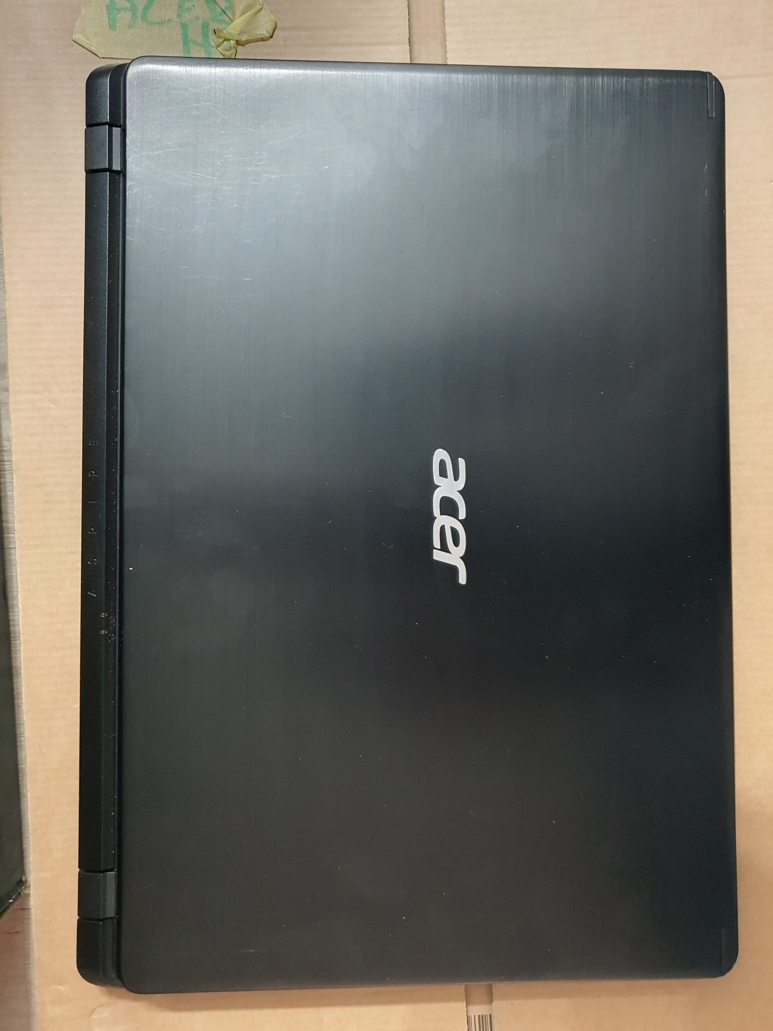 Czarny laptop Acer z widocznym logo, zamknięty, leżący na tekturowym podłożu. Widoczne delikatne rysy na obudowie.