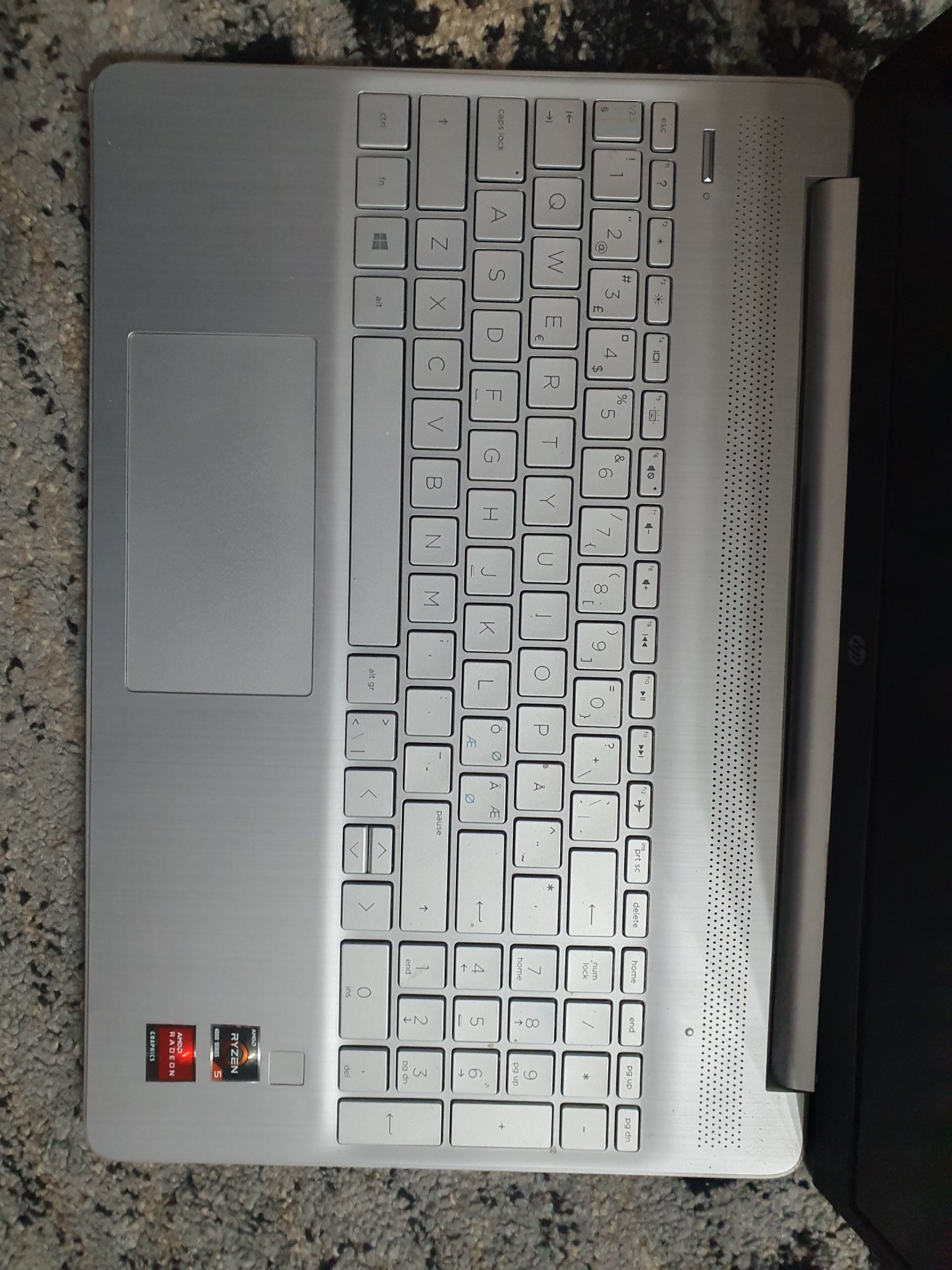 Laptop HP z procesorem AMD Ryzen 5, widoczny z góry. Klawiatura QWERTY, touchpad i naklejki z logo AMD. Szary odcień obudowy. Widoczny fragment dywanu w tle.