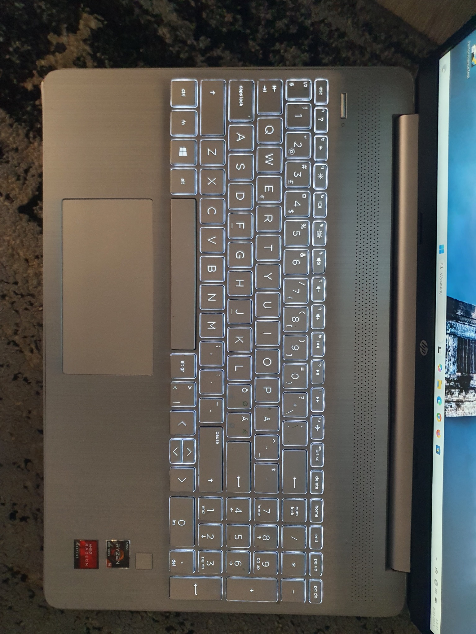 Podświetlana klawiatura laptopa HP z procesorem AMD Ryzen i grafiką Radeon, widoczny ekran z systemem Windows. Laptop leży na ciemnym, wzorzystym tle.