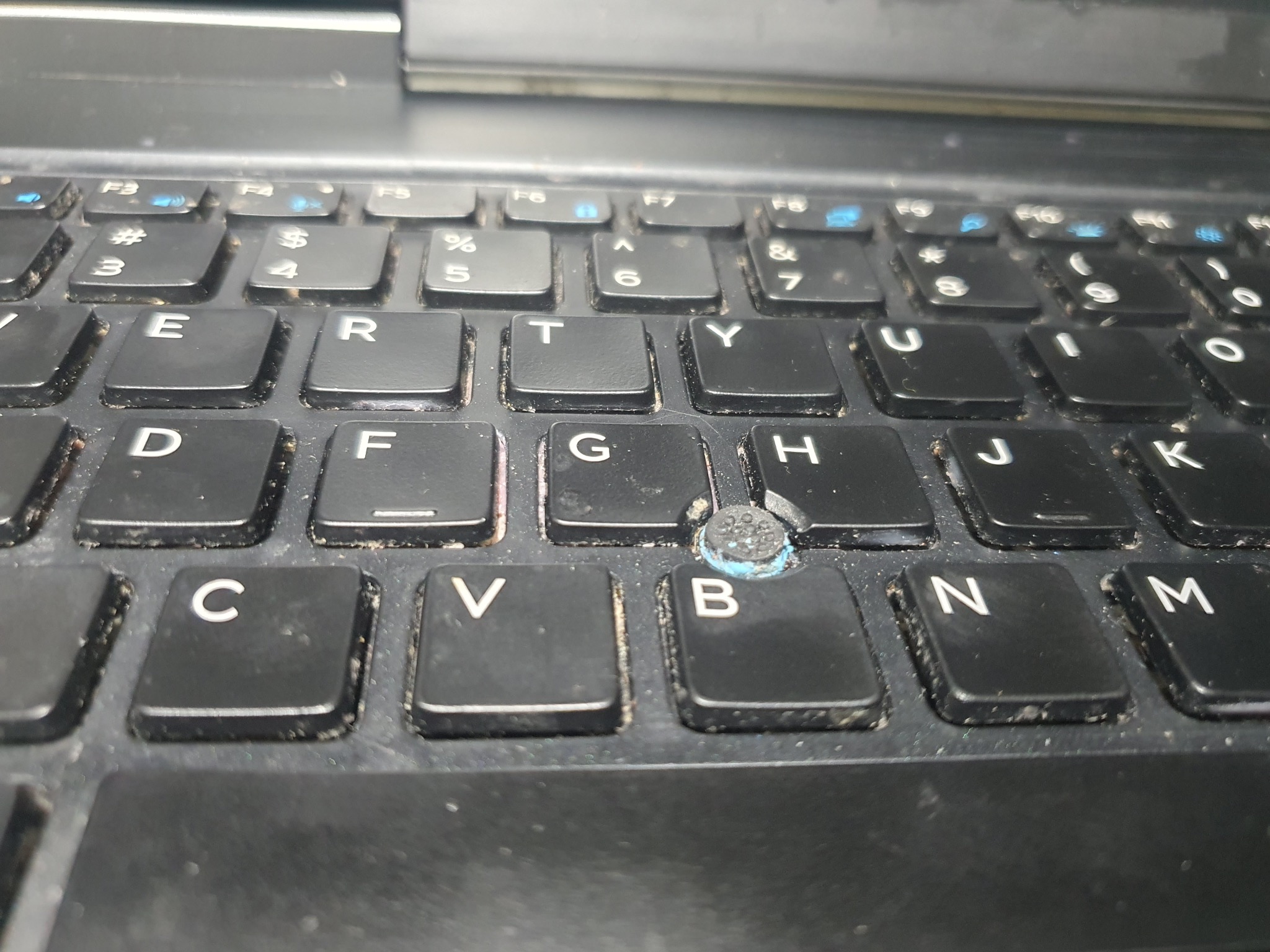 Brudna klawiatura laptopa z bliska, widoczne czarne klawisze z białymi literami i niebieskimi symbolami funkcyjnymi. Klawisz 'G' z uszkodzonym gumowym elementem.
