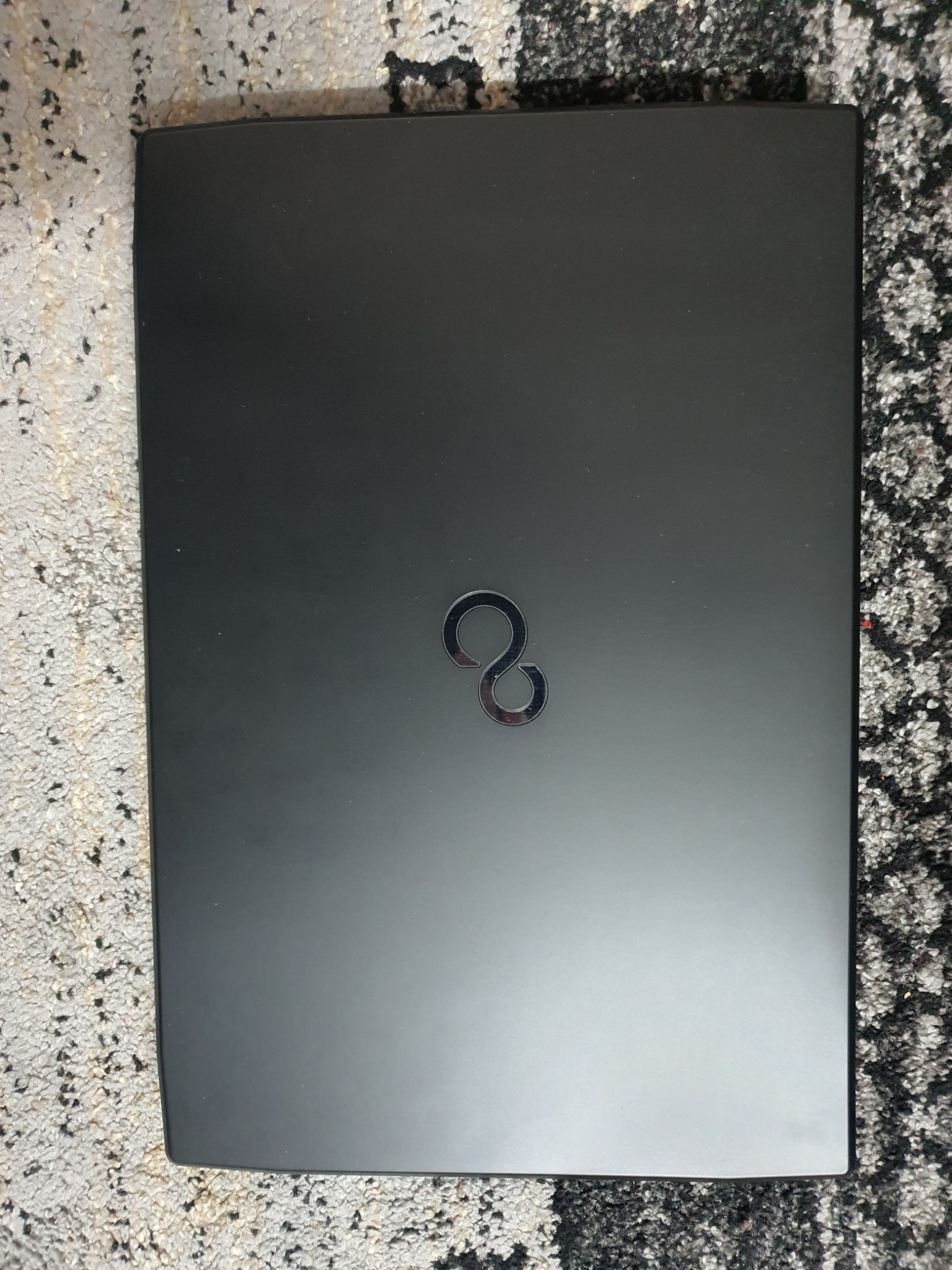 Czarny laptop Fujitsu z metalowym logo na tle dywanu o szaro-czarnej fakturze. Zamknięty laptop leży płasko, widoczny z góry.