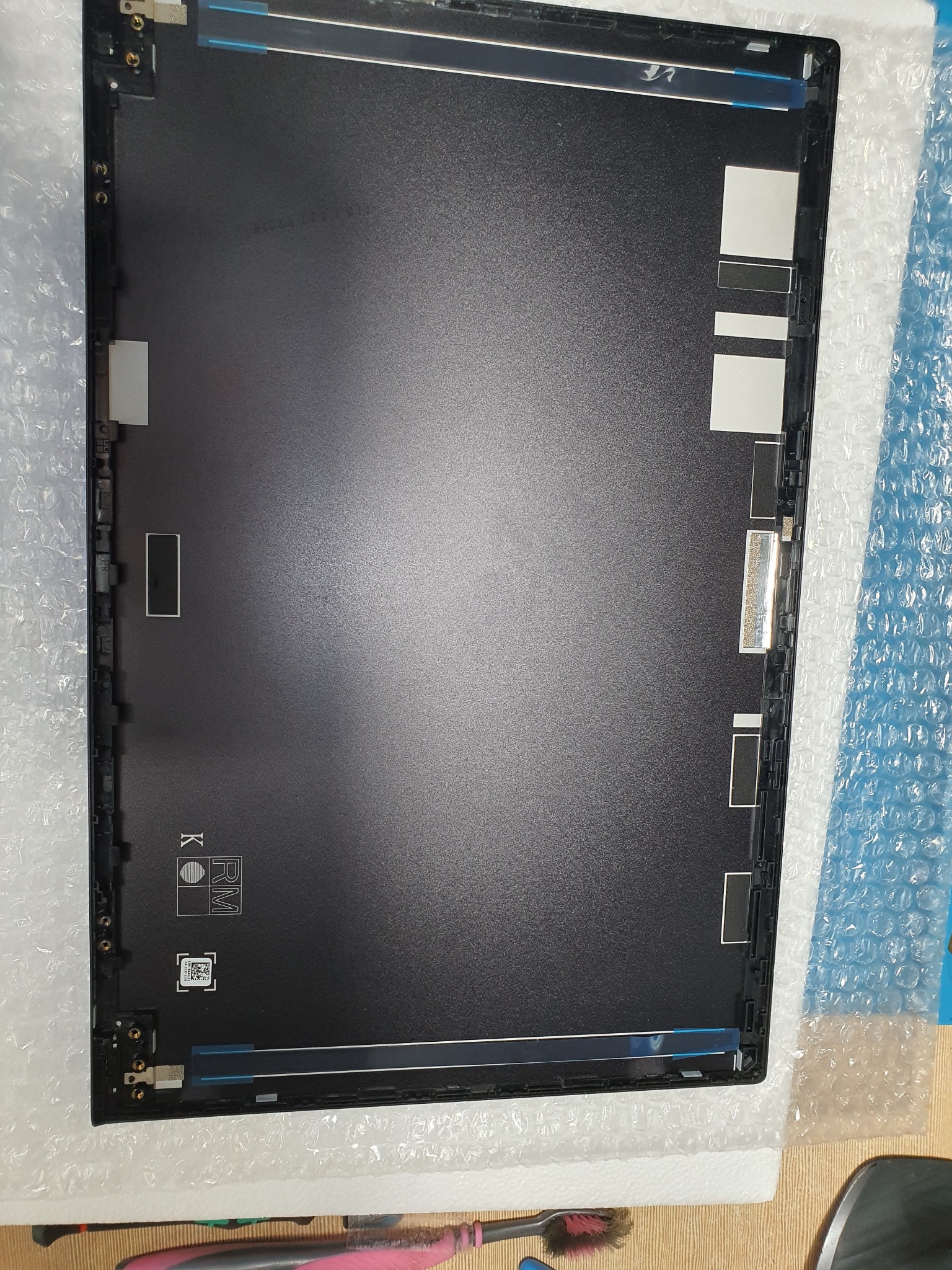 Wnętrze obudowy laptopa z widocznymi elementami mocującymi i taśmami. Czarna powierzchnia, fragment folii bąbelkowej w tle, narzędzie czyszczące w rogu.