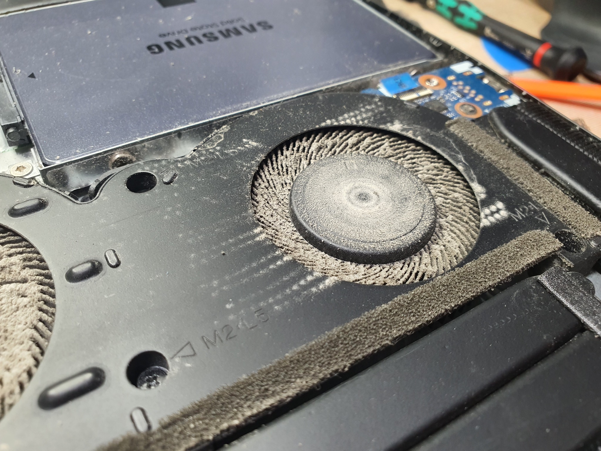 Wnętrze laptopa z zakurzonym wentylatorem i dyskiem SSD Samsung. Widoczne ślady użytkowania i narzędzia serwisowe w tle, sugerujące proces czyszczenia lub naprawy.