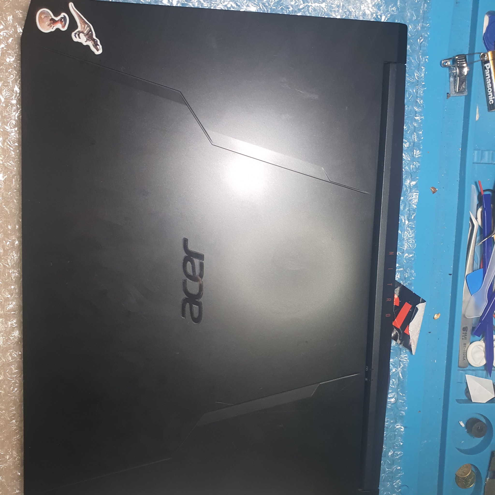 Czarny laptop Acer Nitro z naklejkami na obudowie, leżący na niebieskiej macie serwisowej z narzędziami, sugerujący naprawę lub serwis.