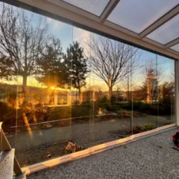 Szklana zabudowa balkonu z widokiem na ogród o zachodzie słońca. Widoczne przesuwne drzwi szklane i metalowa konstrukcja zadaszenia. Częściowo widoczna drabina.